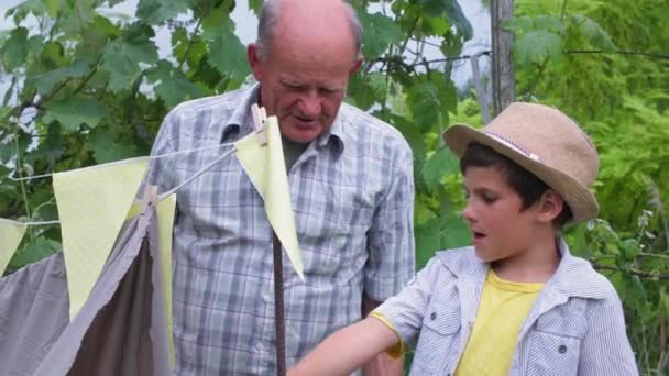 Estilo de vida, pequeño niño masculino sonriente en sombrero con su abuelo divertirse en el fondo del jardín de árboles verdes en el día cálido — Vídeo de stock