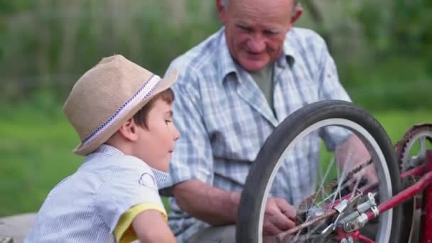 Happy male child with grandfather rejoices at repairing wheel on bicycle and raises his hands up making cheers gesture backdrop of green trees — Stock Video