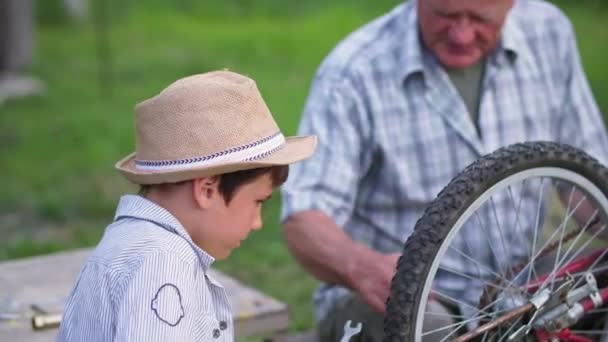 Família amigável, criança masculina diligente, juntamente com um homem idoso, reparos roda de bicicleta com ferramentas durante as férias de verão fora da cidade — Vídeo de Stock