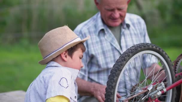 Cheerful male child, together with caring grandfather, repairs bicycle wheel while walking outside city — Stock Video