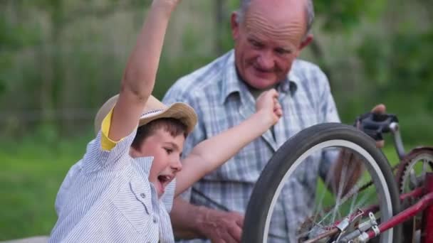 Férias na aldeia, neto feliz com seu avô repara roda de bicicleta enquanto caminha, levanta as mãos fazendo gestos de alegria — Vídeo de Stock