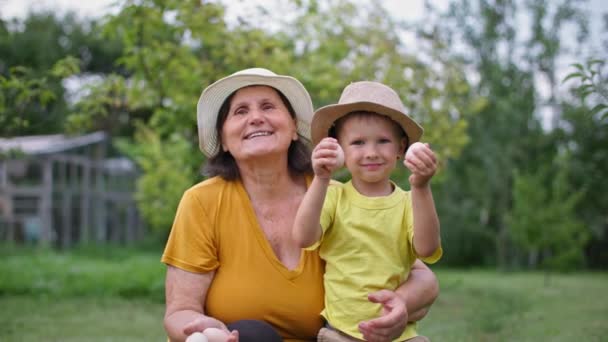 Органічні домашні фермерські яйця, бабуся з онуком в солом'яних капелюхах позує на камеру з яйцями в руках в зеленому саду — стокове відео