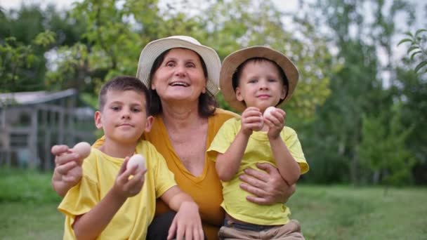 Домашня сімейна куряча ферма бізнес, бабуся з онуками в солом'яних капелюхах позує на камеру з яйцями в руках на задньому дворі — стокове відео