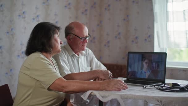 Medicina en línea, pareja casada jubilada con pastillas consultar a una doctora a través de un ordenador portátil sentado en la mesa en casa — Vídeos de Stock