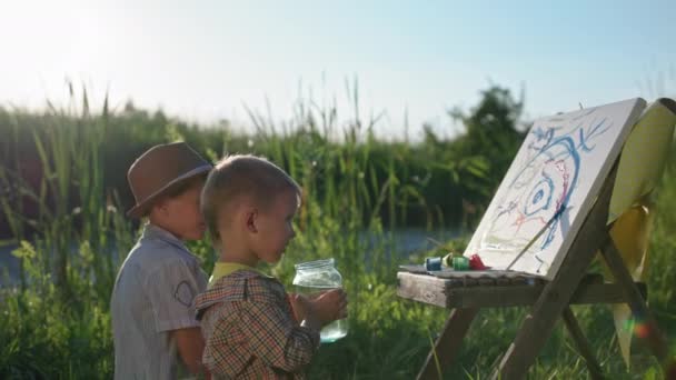 Lifestyle, kleine jongen drinkt water en tekent samen met zijn broer met borstels en verf op papier terwijl ontspannen in de natuur op de achtergrond van groen gras en riet — Stockvideo