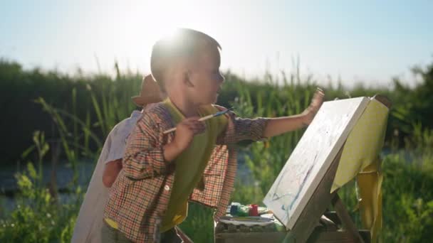 Ontwikkeling van kinderen, kleine creatieve mannelijke kinderen schilderen met verf en penselen op doek tijdens recreatie buitenshuis — Stockvideo