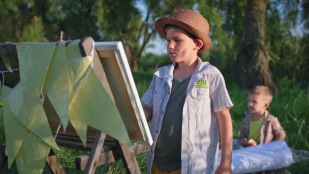 Zorgeloze kindertijd, schattig mannelijk kind in hoed tekent met een borstel en schildert op een ezel met papier, terwijl ontspannen op het platteland, op de achtergrond kleine jongen is het eten van een appel zitten op gras — Stockvideo