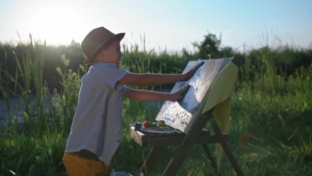 Kinderhobby, slim mannelijk kind in muts tekent met handen en verf op doek met een ezel op achtergrond van groen riet en zonnestralen — Stockvideo