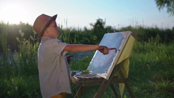 Παιδικό χόμπι, ταλαντούχος μικρός καλλιτέχνης στο καπέλο ζωγραφίζει με τα χέρια και τα χρώματα σε καμβά με ένα καβαλέτο κοντά στο ποτάμι φόντο της δύσης του ήλιου — Αρχείο Βίντεο