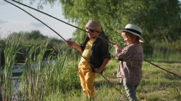 Summer vacation, adorable little boys in hats with fishing rods in their hands are fishing in lake on green lawn among reeds and green trees — Stock Video