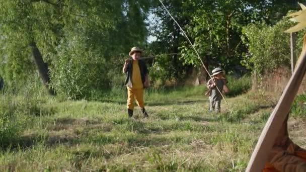 Děti na venkově, roztomilí kluci v kloboucích s rybářskými pruty v rukou se baví relaxaci na trávníku u řeky mezi stromy — Stock video