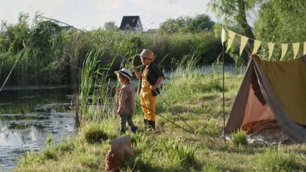 Feliz infancia, niños varones activos en sombreros están pescando en estanques con una caña de pescar durante la recreación al aire libre con wigwam entre árboles y cañas — Vídeos de Stock
