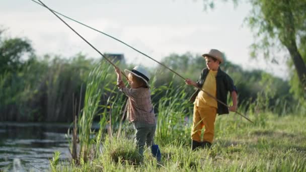 Angler, fröhliche Brüder mit Sonnenhüten fischen mit der Angelrute im Sommerurlaub auf dem Land am Fluss zwischen Bäumen und Schilf — Stockvideo