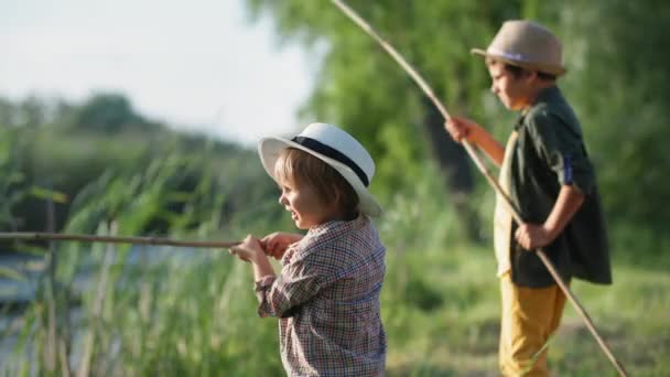 Unterhaltung für Kinder, kleiner männlicher Fischer mit seinem Bruder angelt mit Angelruten im See auf grünem Rasen, während er sich außerhalb der Stadt ausruht — Stockvideo