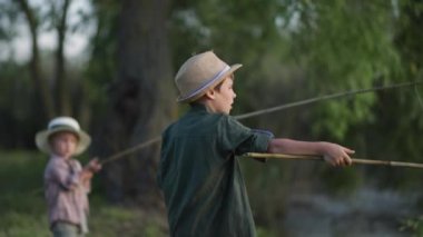 Çocuklar balıkçılar, küçük kardeşiyle şapkalı sevimli bir çocuk nehir kenarında dinlenirken tahta bir çubukla balık tutuyor.