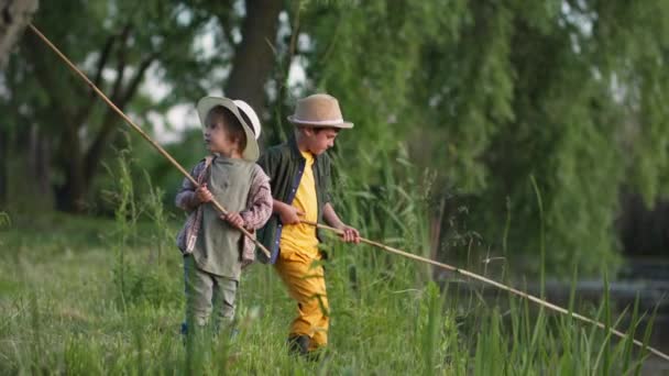 Little fisherman in straw hats with fishing rods in their hands have fun fishing in pond among trees and weeds on warm summer evening while relaxing in countryside — Stock Video