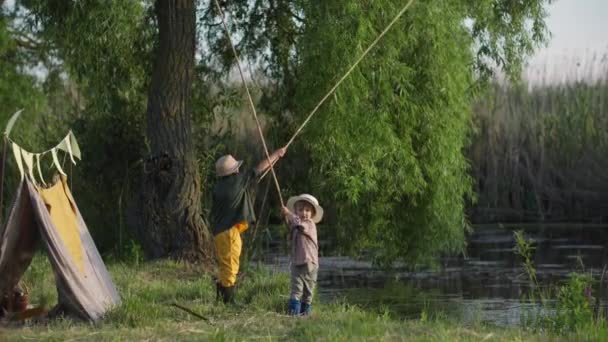 Šťastné děti ve slunečních čepicích s rybářskými pruty v rukou chytají ryby v jezeře během letních prázdnin na venkově — Stock video