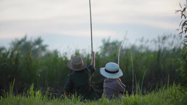 Niños al aire libre, niños alegres en sombreros de paja con cañas de pescar en sus manos atrapar peces en la orilla del río sentado en el prado entre cañas al atardecer — Vídeos de Stock
