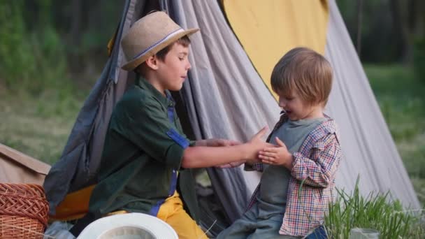 Omtänksamt manligt barn leker sina händer med sin yngre bror när han sitter i wigwam under sommarlovet utanför staden — Stockvideo
