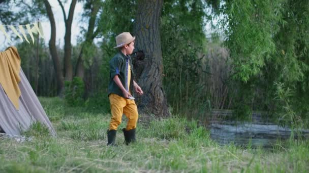 Fin de semana de verano, niño varón activo en botas de goma y sombrero se divierte tirando piedras al río mientras se relaja fuera de la ciudad — Vídeos de Stock