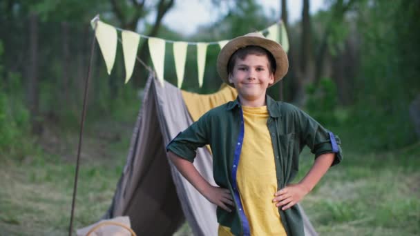 Портрет усміхненої чоловічої дитини в капелюсі просякнутий фоном відпочинку на відкритому повітрі вігвама в сільській місцевості — стокове відео