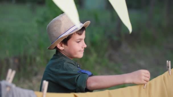 Rozkošný chlapec v klobouku dělá wigwam a visí kolíky oblečení při relaxaci venku na letním večeru na venkově — Stock video
