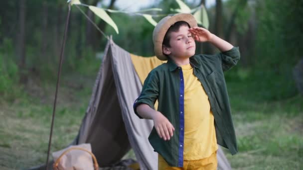 Portrait of joyful boy in hat has fun on vacation outdoors and shows thumbs up background of wigwam and trees, look at camera — Stock Video