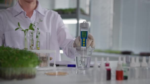 Laboratorios de plantas, asistente de laboratorio femenino examina la calidad del agua con un dispositivo de medición para regar los brotes de microverdes jóvenes en el moderno telón de fondo de invernadero de los estantes, primer plano — Vídeo de stock