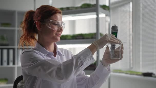 O laboratório de engenharia genética da pesquisa biológica, trabalhador do laboratório fêmea nos óculos protetores conduz a pesquisa com água para irrigar micro-verdes e goteja o reagente no fundo do frasco de — Vídeo de Stock