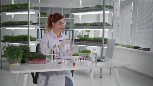 Retrato de bióloga cientista conversando por videoconferência em laptop e discutindo planta em tubo de ensaio com auxiliares de laboratório sentados em laboratório, sorrindo e olhando para a câmera — Vídeo de Stock