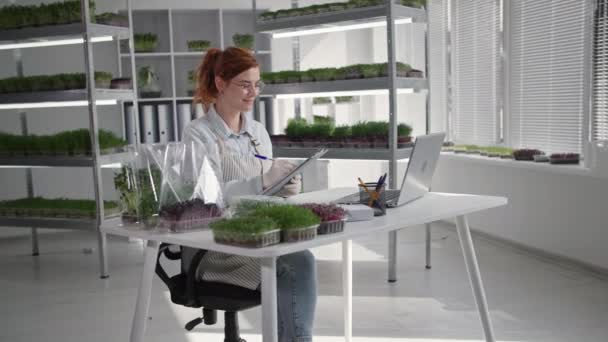 Νεαρή γυναίκα πωλητής εργάζεται σε μικρο αγρόκτημα πρασίνου και πωλεί την ανάπτυξη συσκευασίας σε απευθείας σύνδεση μέσω της επικοινωνίας βίντεο στο laptop κάθεται στο φόντο τραπέζι των ραφιών — Αρχείο Βίντεο