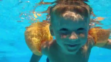 happy kid boy with open eyes swims under the water in blue swimming pool, underwater shot of child with yellow inflatable oversleeves