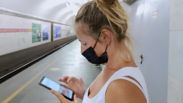 Technology and city life, young woman in protective mask at the metro station uses mobile phone with internet while waiting for transport — ストック動画