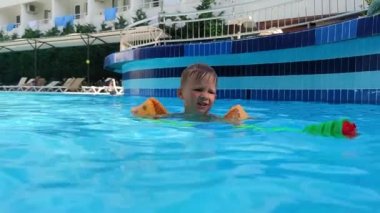 Havuzda sarı şişirilebilir kolları olan küçük çocuk, su tabancası olan çocuk sıcak bir günde kameraya ateş ediyor.