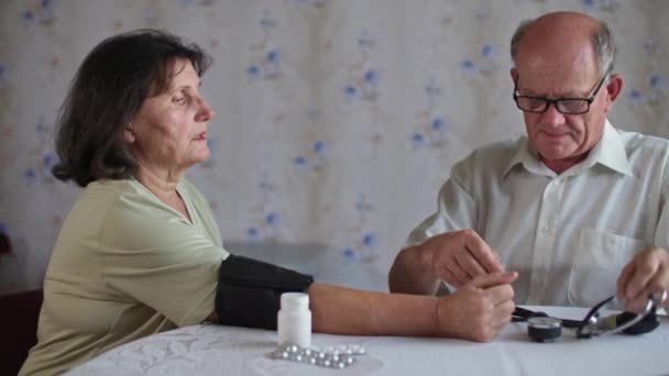 Salud jubilación, cuidado anciano con gafas mide la presión de su esposa con tonómetro sentado en la mesa con pastillas en casa — Vídeo de stock