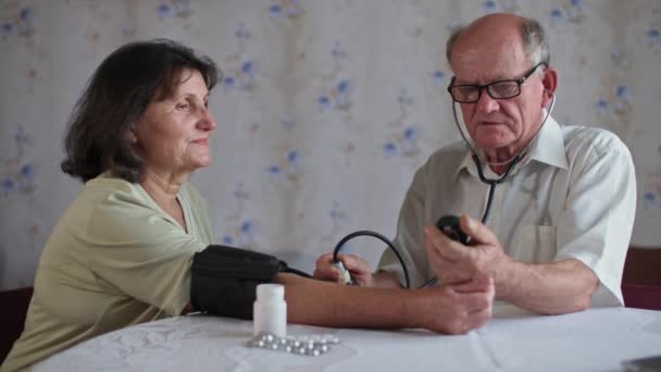 Medición de la presión y el pulso, el hombre de edad avanzada utiliza un tonómetro mientras está sentado junto a su esposa en casa — Vídeo de stock