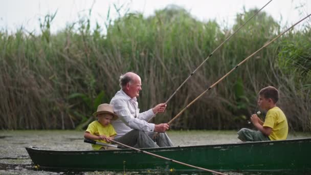 Семейные выходные на пруду, рыбак надевает очки, чтобы распутать удочки, сидя в лодке со своими внуками — стоковое видео