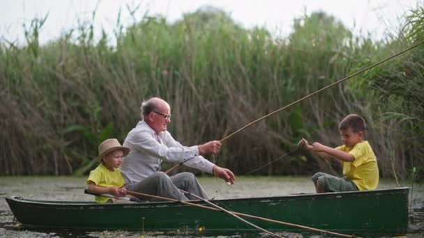 釣りをしながら親のいる小さな男の子、眼鏡をかけた祖父は葦の近くの池にボートに座っている間に孫釣り棒を与えます — ストック動画