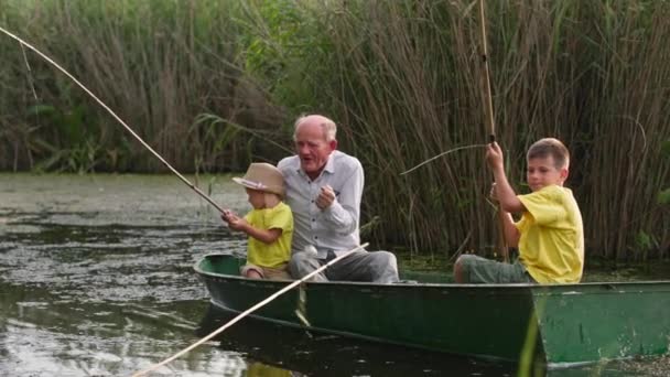 Niños pescando con cañas, abuelo enseña a pescar mientras está sentado en barco con sus nietos en el fondo de cañas durante unas vacaciones de verano — Vídeos de Stock