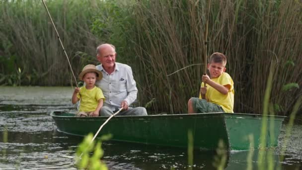 Miły dziadek uczy swoje wnuki łowić ryby na rzece, rodzina bawiąc się razem w łodzi w pobliżu trzciny na letnie wakacje na wsi — Wideo stockowe