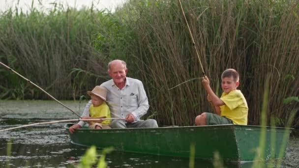 Активное детство на реке, дед с внуками рыбачат, сидя в лодке на фоне тростника — стоковое видео