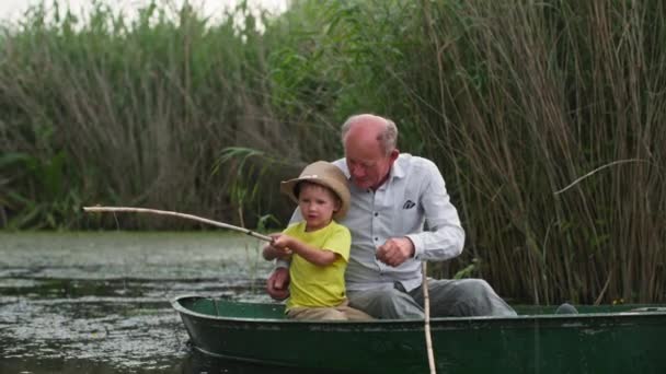 Childhood, little cute male child together with his beloved grandfather in boat, fisherman teaches his grandson to fish in a pond with fishing rod — Stock Video