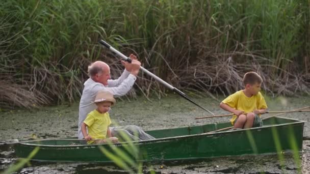 Дедушка катается на своих внуках на лодке с веслом на фоне тростника, семейный отдых во время рыбалки на пруду — стоковое видео