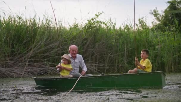 Happy childhood, grandfather teaches grandchildren to fish using fishing rods while sitting in boat on background of reeds — Stock Video