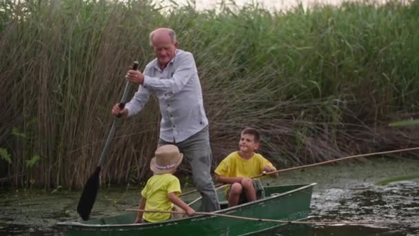 Padre mayor de pie en el barco remando un remo a lo largo del río, abuelo rueda a sus nietos en el estanque mientras pesca — Vídeos de Stock