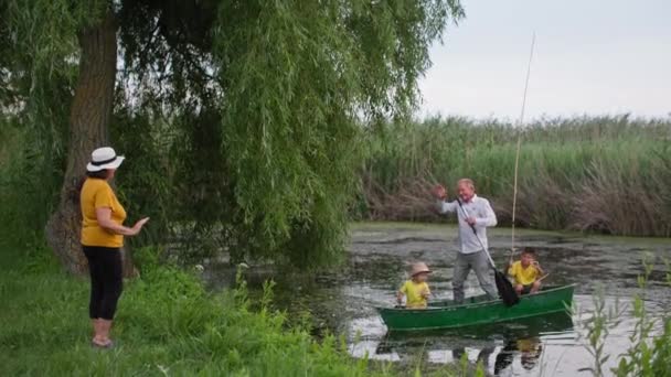 Бабушка приветствует внуков с рыбалки, старших родителей гребное весло, стоя на лодке с маленькими пассажирами на фоне красивой природы — стоковое видео