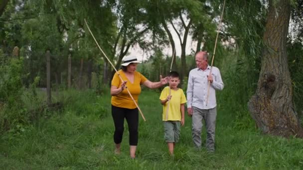 Familia feliz hablando de la pesca mientras camina en el campo, los abuelos van a lo largo del césped verde con el nieto sosteniendo palos en las manos — Vídeos de Stock