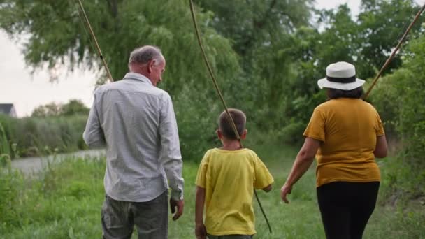 Vista posteriore la famiglia sta camminando in campagna con canne da pesca in mano, nipotino racconta ai nonni di pesca vicino al fiume — Video Stock