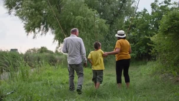 Abrazo familiar, los abuelos están caminando por el césped verde con su nieto pequeño con cañas de pescar en sus manos, vista trasera — Vídeos de Stock