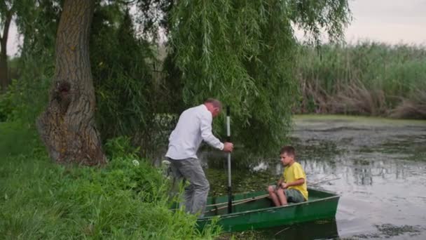 Семья собирается на рыбалку, дедушка и внук садятся в лодку и плывут вдоль реки с помощью весел — стоковое видео